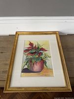 Frontal view of the signed watercolour painting featuring a potted plant with red and green foliage in a gold frame on a wooden surface.