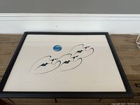 Framed lithograph artwork featuring abstract blue and black forms with a blue circle to the upper left, laying flat on wood surface.