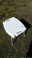 White plastic seat of InnaCare bath stool outdoors on grass, showing shape, handles and drainage indentations.