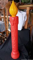 Tall red Christmas candle blow mold with "NOEL" embossed, yellow flame light top, standing on a black cloth.