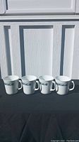 Front view of four CorningWare Old Town mugs with blue and green leaf pattern, arranged in a row on black cloth against white panel background.