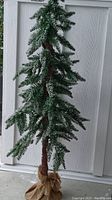 Artificial pine tree about 3 feet tall, with snow-like flocking on the needles and branches, set against a textured white door background.