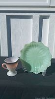 Front view of a green Jadeite plate with scalloped edges and a short pedestal pink to black creamer placed on black cloth against a white wood panel background