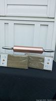 Metal rolling pin displayed horizontally on top of two sets of napkins with green and beige checked pattern in original sealed packaging.