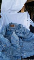 Light wash stretch jean jacket displayed on a chair with white cloth background, showing front and sleeves details.