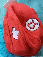 Red fleece hockey helmet style hat with embroidered maple leaf and Lululemon logos and adjustable chin strap, shown on textured turquoise background.