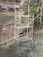 Photo showing a white metal folding shelf with decorative scrollwork, white metal magazine holder, five black metal plant hangers, and a black metal plant stand on outdoor gravel surface.