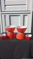 Front view of 2 red Pyrex bowls placed inverted and 2 small rectangular fridge keeper containers in red with white interiors on a black cloth against a white wood background.