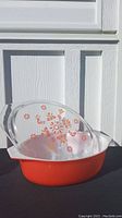 Front and angled view of vintage large oval Pyrex casserole dish on black surface with white paneled background. Dish is red-orange with clear lid featuring orange floral and bird pattern.