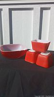 Photo shows set of four red Pyrex dishes: one large square serving bowl and three smaller square refrigerator dishes arranged in sunlight on black cloth with white wall backdrop.