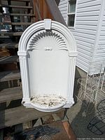 Front view of white resin decorative niche showing arched scalloped top, vertical ridges on sides, and curved base with dirt requiring cleaning.