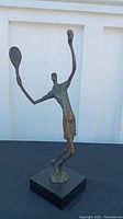 Tennis player sculpture holding two rackets with raised arms on black base.