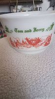 Close-up side view showing green 'Tom and Jerry' text and red carriage and town illustrations on white milk glass bowl.