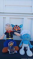 Four vintage plush toys displayed outdoors including Popeye, Furby, Smurf, and Lego Movie character
