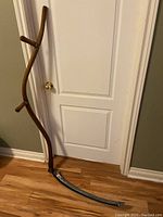 Full view of antique hay scythe leaning against a door, showing entire length including blade and wooden handle.