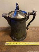Full view of the antique silver plated pitcher with lid closed, showcasing the figurine on the lid and measuring tape for size reference.