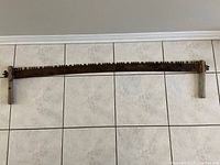 Full view of antique two man crosscut saw horizontally laid on tiled floor showing entire saw including blade and handles.