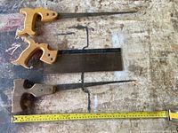 Three small hand saws laid out on a workbench surface with visible measuring tape showing sizes, highlighting the repaired handle on one keyhole saw and the backsaw with a square blade.
