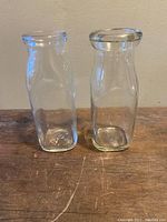 Front view of two small clear glass milk bottles standing upright on a wooden surface against a plain wall.