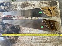 Two vintage large hand saws laid side by side showing full length, measuring tape below for scale.