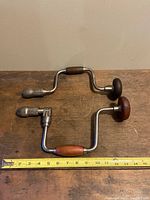 Top view of both vintage brace bit manual hand drills showing the full length of the tools alongside a measuring tape indicating about 14.5 and 13.5 inches respectively. The wood handles and metal frames are visible.