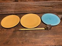 Front view of three Homer Laughlin Fiesta Ware plates, two mustard yellow and one turquoise, with a measuring tape showing sizes.