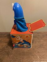 Front view of the vintage Flipper Jack In The Box toy with the dolphin character emerging from the top orange tin box. Detailed lithograph of boy and dolphin on the front panel of the box.