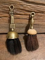 Two vintage horse hair brushes standing upright on the wood surface, showing brass and wood handles with distinct shapes and a Canterbury Cross emblem on the larger brush.