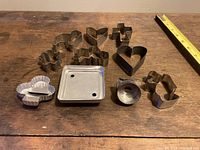 Photo of 10 metal cookie cutters and square container with some wear and vintage patina, showing their shapes and condition on wooden surface with ruler.