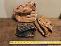 Three vintage baseball gloves lined up on a wooden surface with measuring tape showing approximately 11-12 inch length