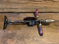 Side view of the vintage Master Craft 107 brace drill showing the black shoulder plate, red gear wheel, and wooden handles.