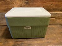 Front and side view of the vintage Thermos plastic cooler showing the textured green body, beige top and base, and branded metal plate on front.