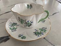 Photo of the Shelley porcelain teacup and matching saucer showing scalloped gold gilded rims and floral pattern.