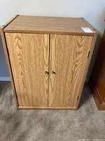 Front view of particle board cabinet with two doors and metal knob handles, wood grain finish.