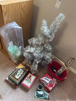 Silver tinsel 32-inch tabletop Christmas tree standing beside a red basket filled with assorted Christmas cards, candles, ornaments, and ornament hooks.