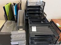 Photo showing black plastic multi-tier letter trays, metal mesh organizers and clear clipboards on desk.