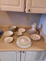Overall view of the dishware lot on counter including stacked plates, bowls, side plates, and mugs.