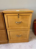 Front view of two-drawer wooden file cabinet in light wood finish, showing metal handles and key in lock.