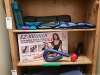 Shelves with EZ-Krunch box and the EZ-Krunch device on display alongside ankle and wrist weights in blue and some dumbbells and a foam mat on upper shelf.