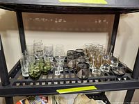 Photo showing various glasses arranged on a black shelf including green tinted glasses, plain clear glasses, and decorative glasses with gold and silver colored patterns.