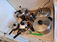 Photo of multiple stainless steel pots and pans including a pressure cooker pot with lids and black handles.