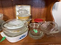 Various Pyrex dishes in different sizes and colors with lids displayed on a wood shelf along with a framed print The Kitchen Prayer