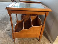 Side view showing wood grain, turned legs, beveled glass top and three-section magazine rack.