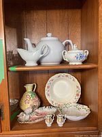 Two wooden shelves containing various ceramic and porcelain kitchenware including a white teapot, gravy boat, decorative sugar bowl, floral plate, grape cluster pitcher, leaf-shaped dish, and salt and pepper shakers.