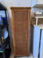Front view of tall wicker cabinet with narrow door and round wooden knob, next to kitchen counters and shelves.