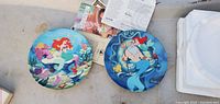 Top view of two round Disney collector plates, one showing Ariel the mermaid and the other depicting Poseidon, both with colorful underwater illustrations.