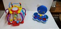 Two electronic learning toys placed side by side on a white surface: a blue octopus toy with interactive buttons and a colorful bead maze cube with various buttons and wire bead activity.