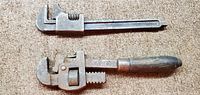 Photo showing two vintage 8-inch pipe wrenches on carpet, one with a metal handle and one with a wooden handle, both with adjustable jaws.