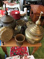 Brass urns and containers arranged on wooden table with some in background including red plates.