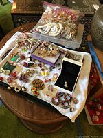 Full overview of costume jewelry items laid out on white towel including earrings, pins, necklaces, and a plastic bin of loose jewelry in background.